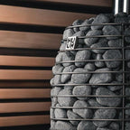 Huum Hive Flow 9.8kW wood-burning sauna stove with sauna stones beside a slatted wood sauna wall
