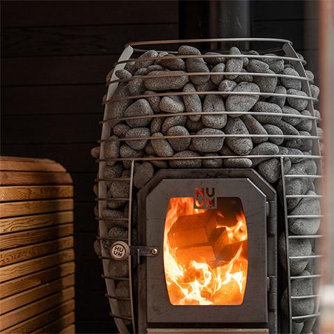 Huum Hive Wood 13kW wood-burning sauna stove burning in a sauna with wooden benches and stacked sauna stones