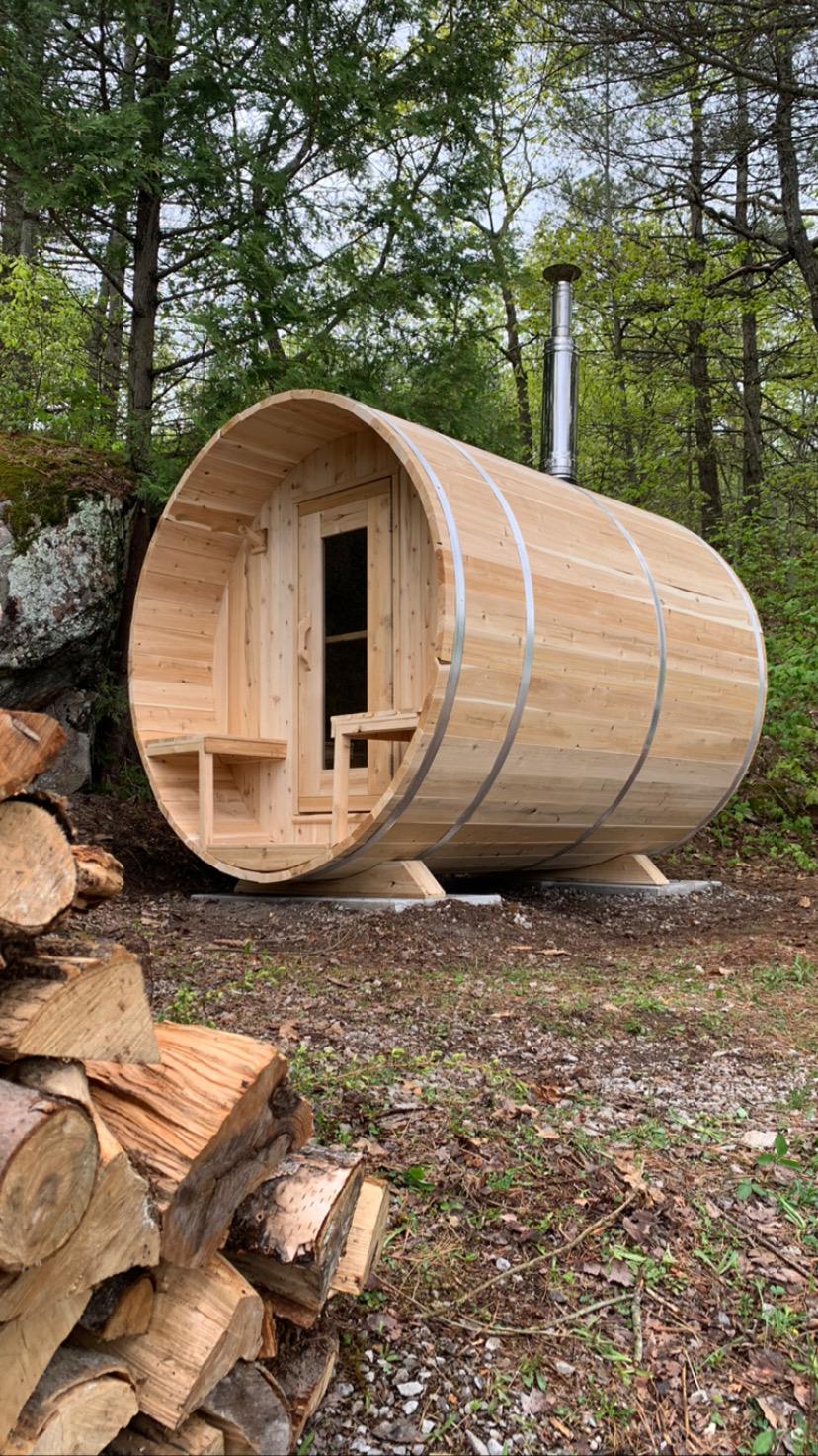 Dundalk CT Serenity 5-Person Outdoor Barrel Sauna beside stacked firewood