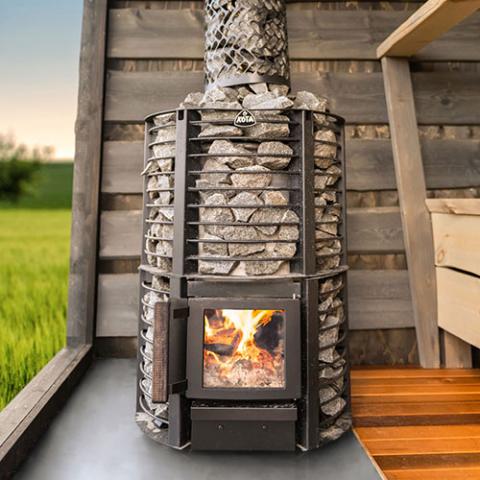 Narvi Inari Plus 16kW wood burning sauna stove with fire burning and a stone-covered chimney in an outdoor sauna