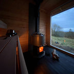 Narvi Luosto VS 16kW wood burning sauna stove with fire burning in a log-walled sauna beside a window